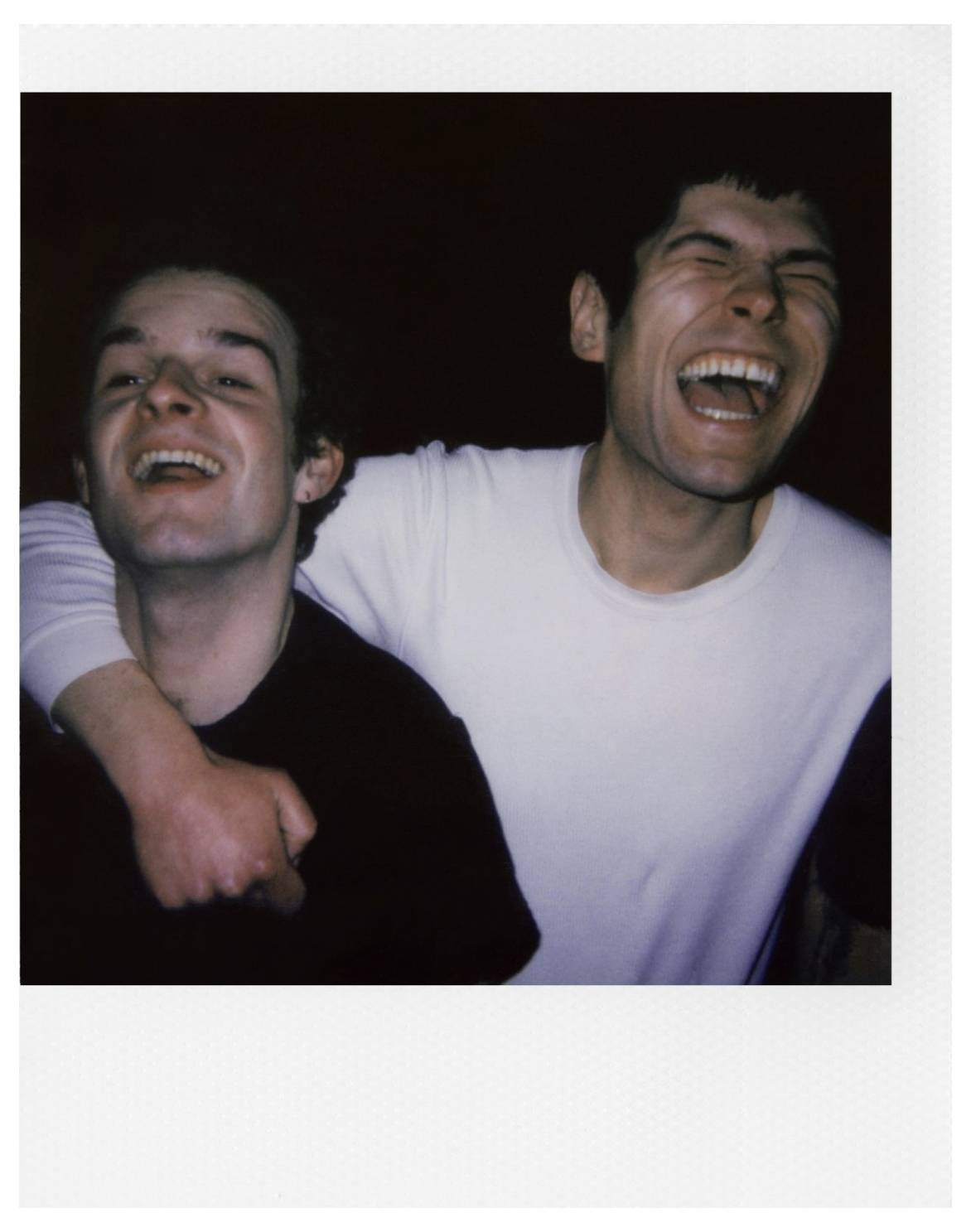 Two people are laughing, one in a black shirt with their arm around the other in a white shirt. The setting is dimly lit.
