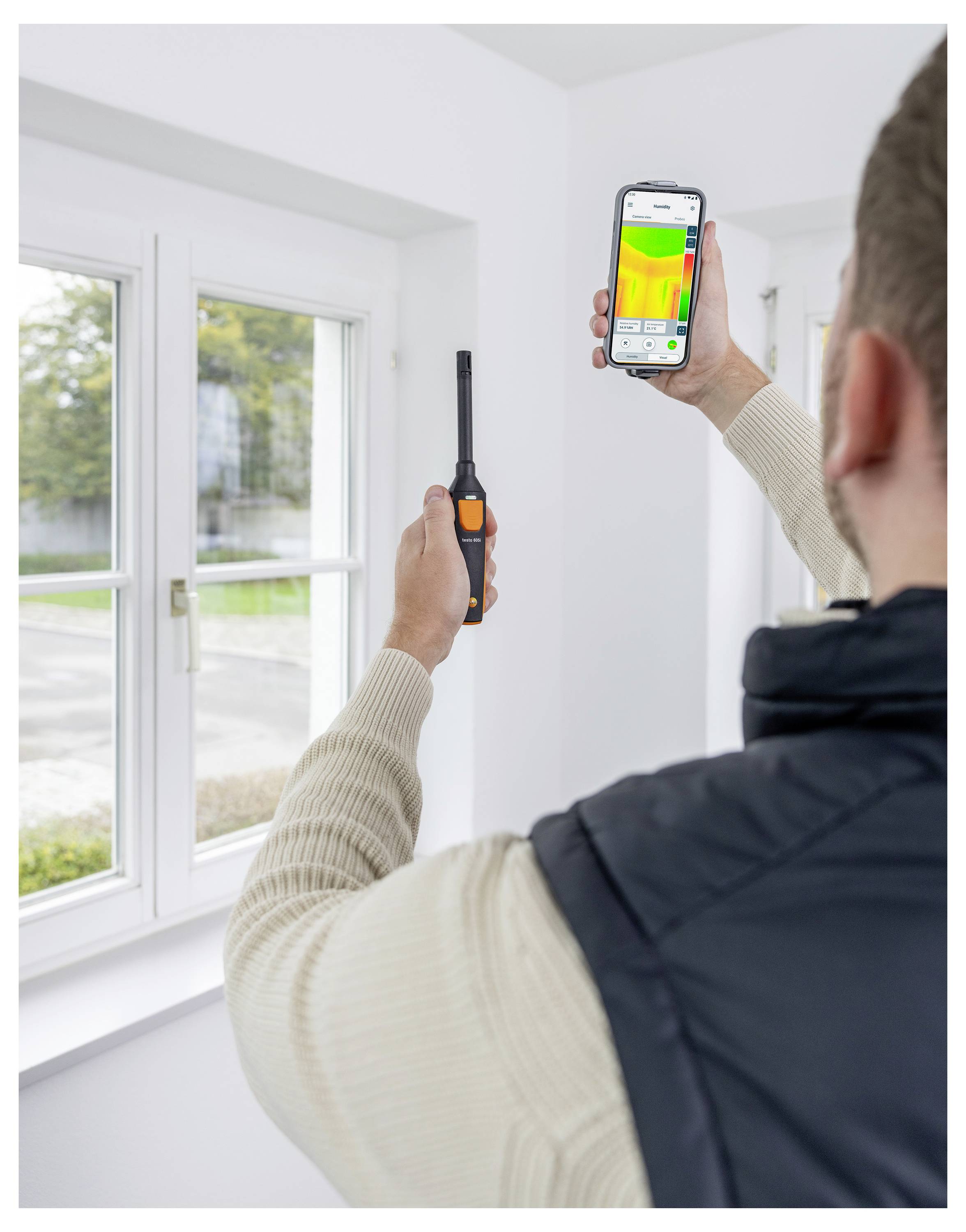 'A person uses a smartphone with an attached thermal imaging device to scan the wall near a window, displaying a colorful heat map.'