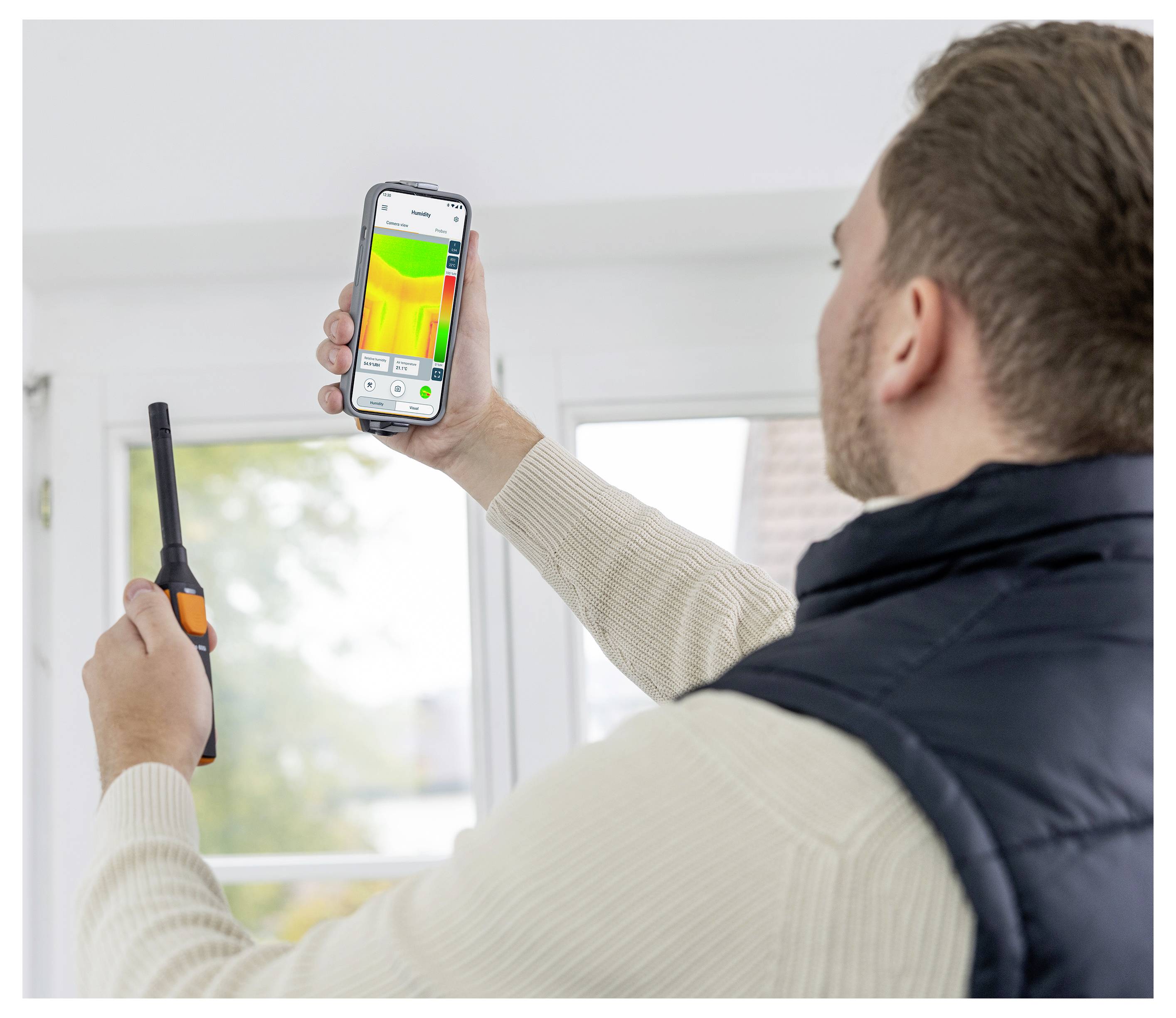 A person holds a smartphone displaying a thermal image app while using a handheld device. They appear to be checking for temperature variations indoors.