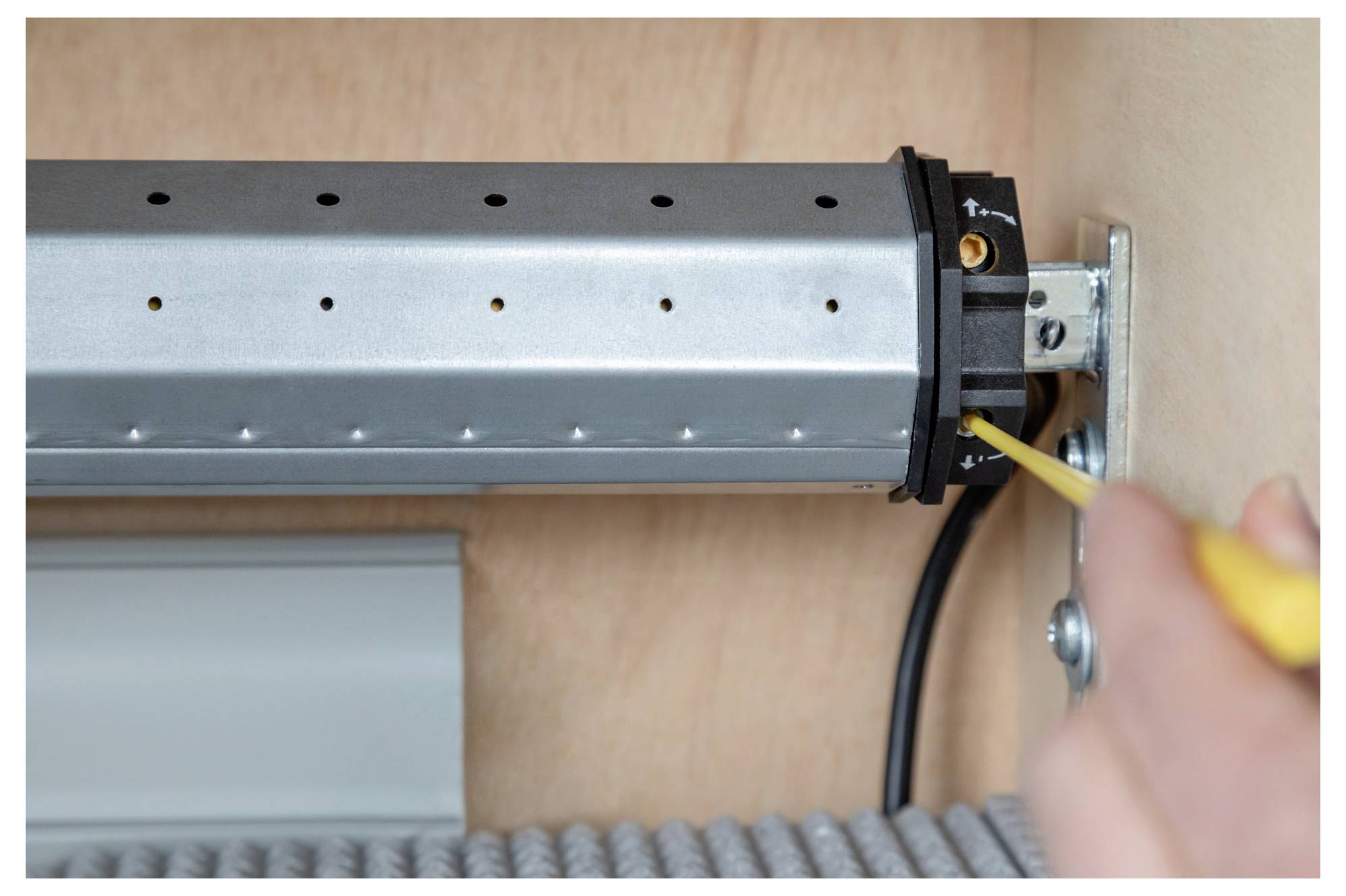 A person tightens a screw on a metal tube with a screwdriver, part of a mechanical assembly, against a wooden background.
