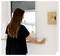 A woman in a black shirt presses a button on a wall switch under a window. A wooden key holder labeled 'KEYS' with a keyhole is nearby.