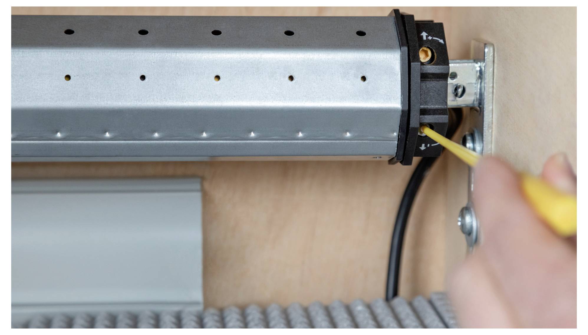 A close-up of a person using a screwdriver to adjust a metal mechanism, possibly part of a roller shutter system, on a wooden surface.