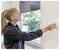 A person with a ponytail is adjusting a thermostat on a wall beside a window with a view of trees outside.