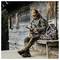 A person in camouflage clothing and boots sits on a wooden bench by a rustic cabin, holding a small tool, with a matching backpack beside them.