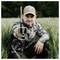 A person wearing camouflage and a cap is kneeling in a field, holding a camouflage trail camera towards the viewer.
