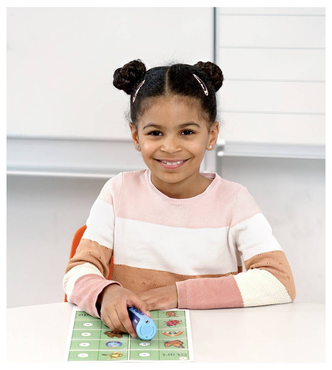 A smiling child with braided hair sits at a desk holding a blue pen-like object over a colorful learning board.
