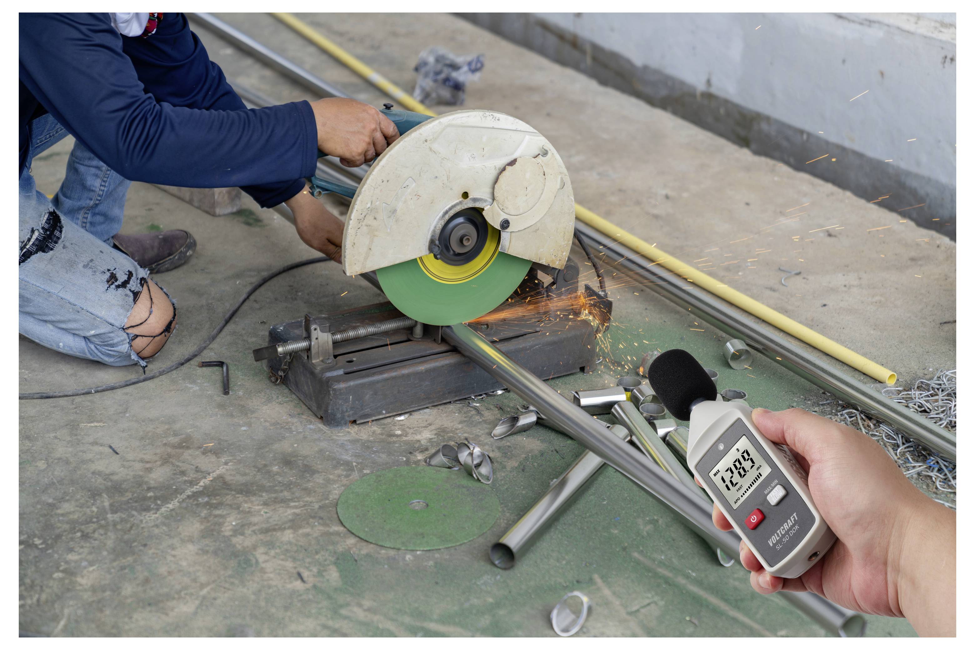 Worker cutting a metal pipe with an electric saw while another person measures the noise level with a handheld decibel meter.