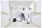 A person vacuuming a hallway with white walls and doors, using a black vacuum cleaner. The floor is beige, and there are handrails on the walls.