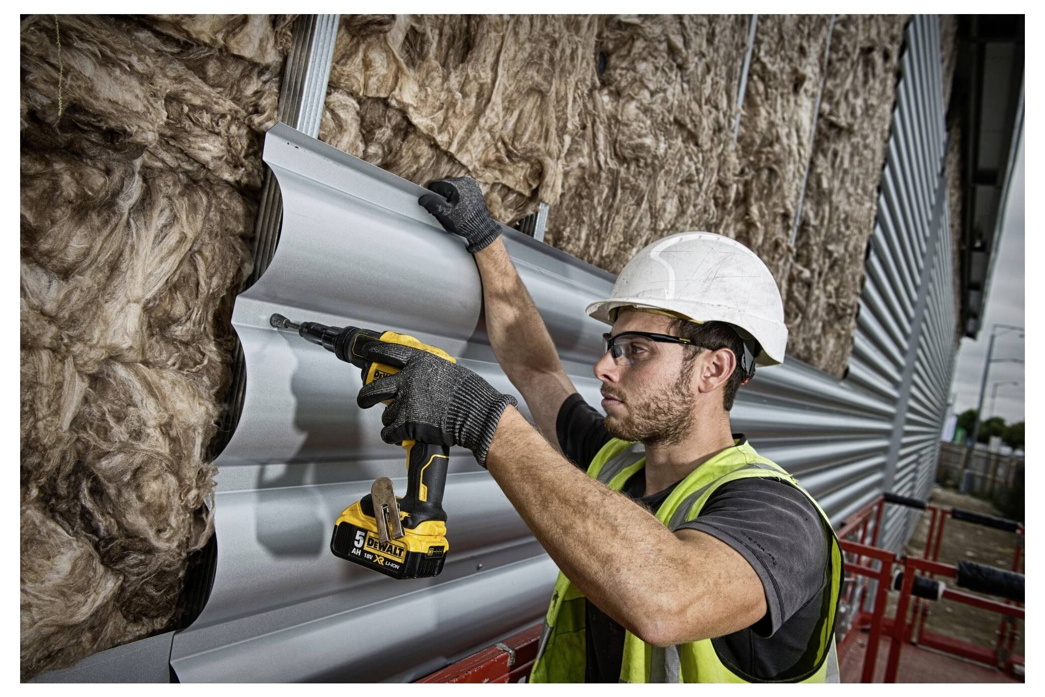 A construction worker in a safety vest and helmet uses a screwdriver to attach corrugated metal siding to a building insulated with fiberglass.