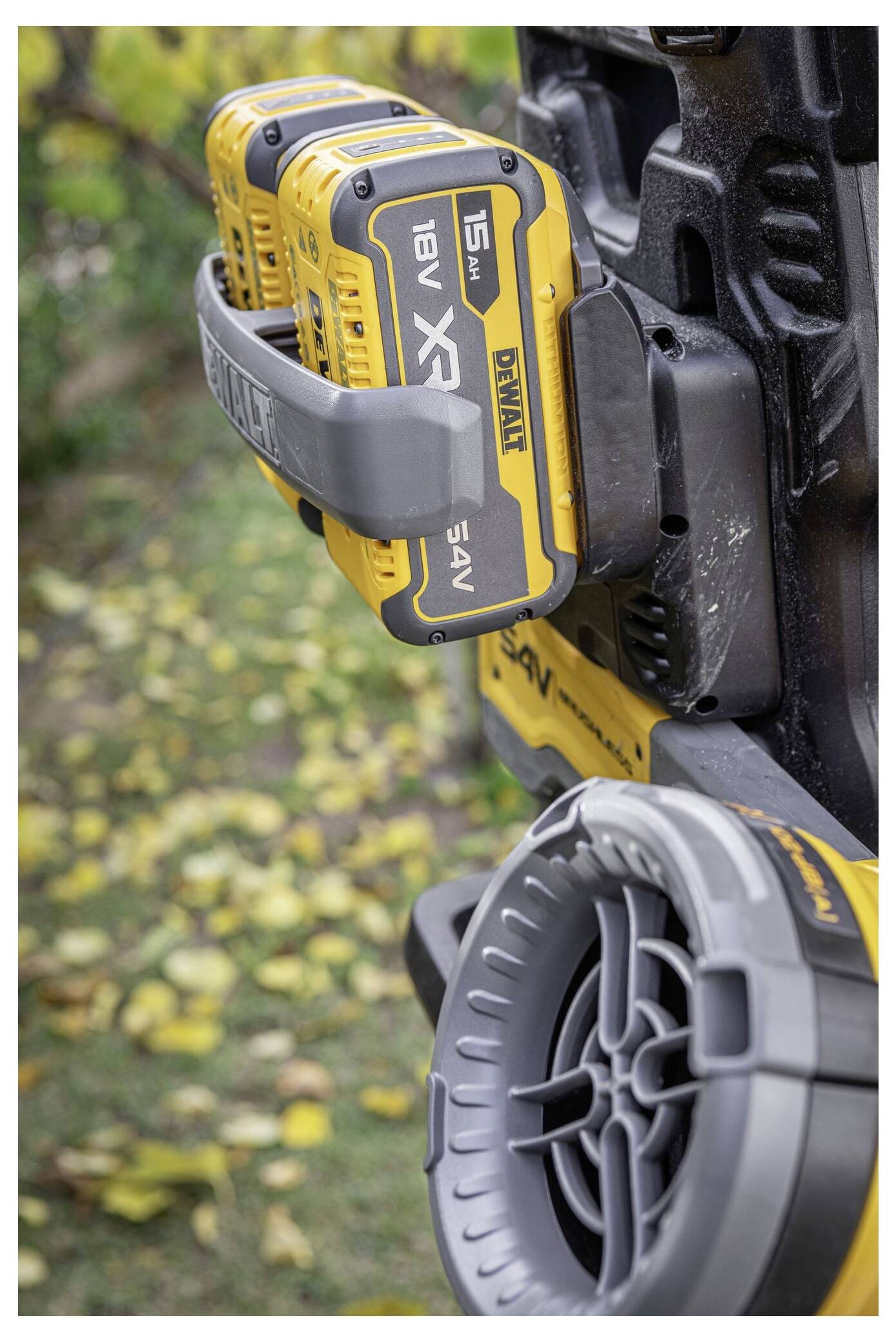 A power tool battery unit with multiple yellow batteries attached, featuring brand logos. In the background, yellow leaves on the ground.
