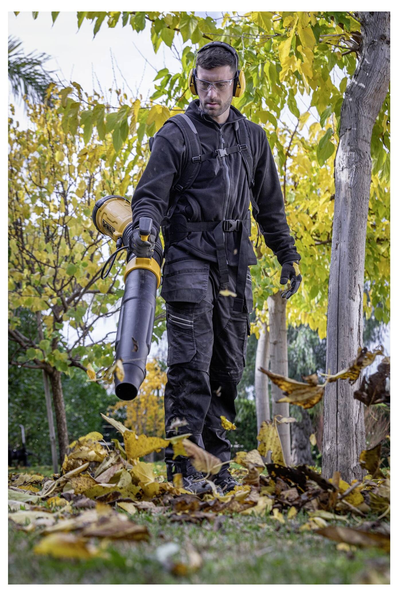 A person in protective gear uses a leaf blower to clear fallen leaves from the ground in a park with trees and autumn foliage.