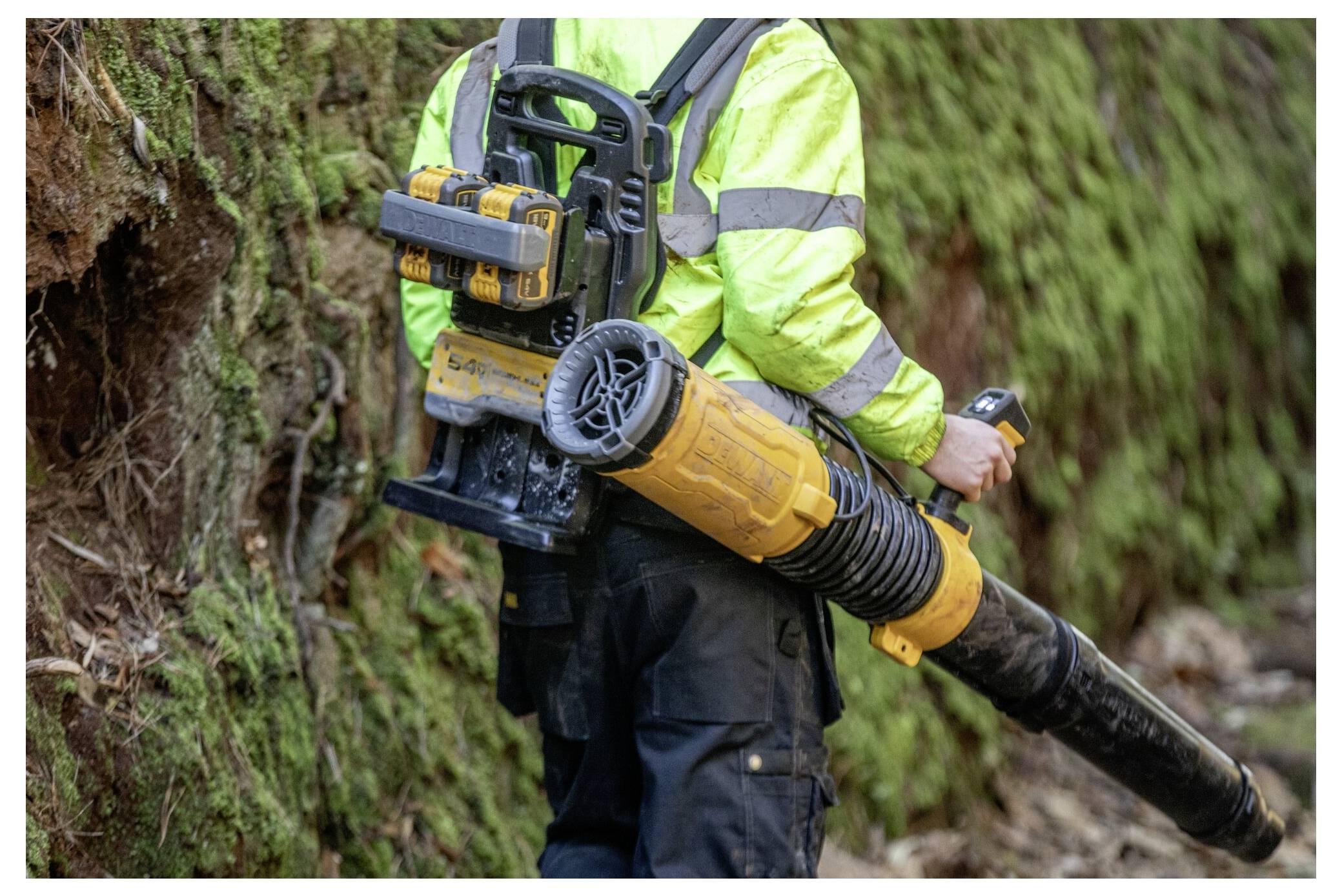 A worker in high-visibility gear carries a yellow and black leaf blower in a forested area with dense moss on the ground.