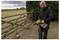 A person in protective eyewear uses a power washer to clean a shovel outdoors, next to a wooden fence and hedge, on a cloudy day.