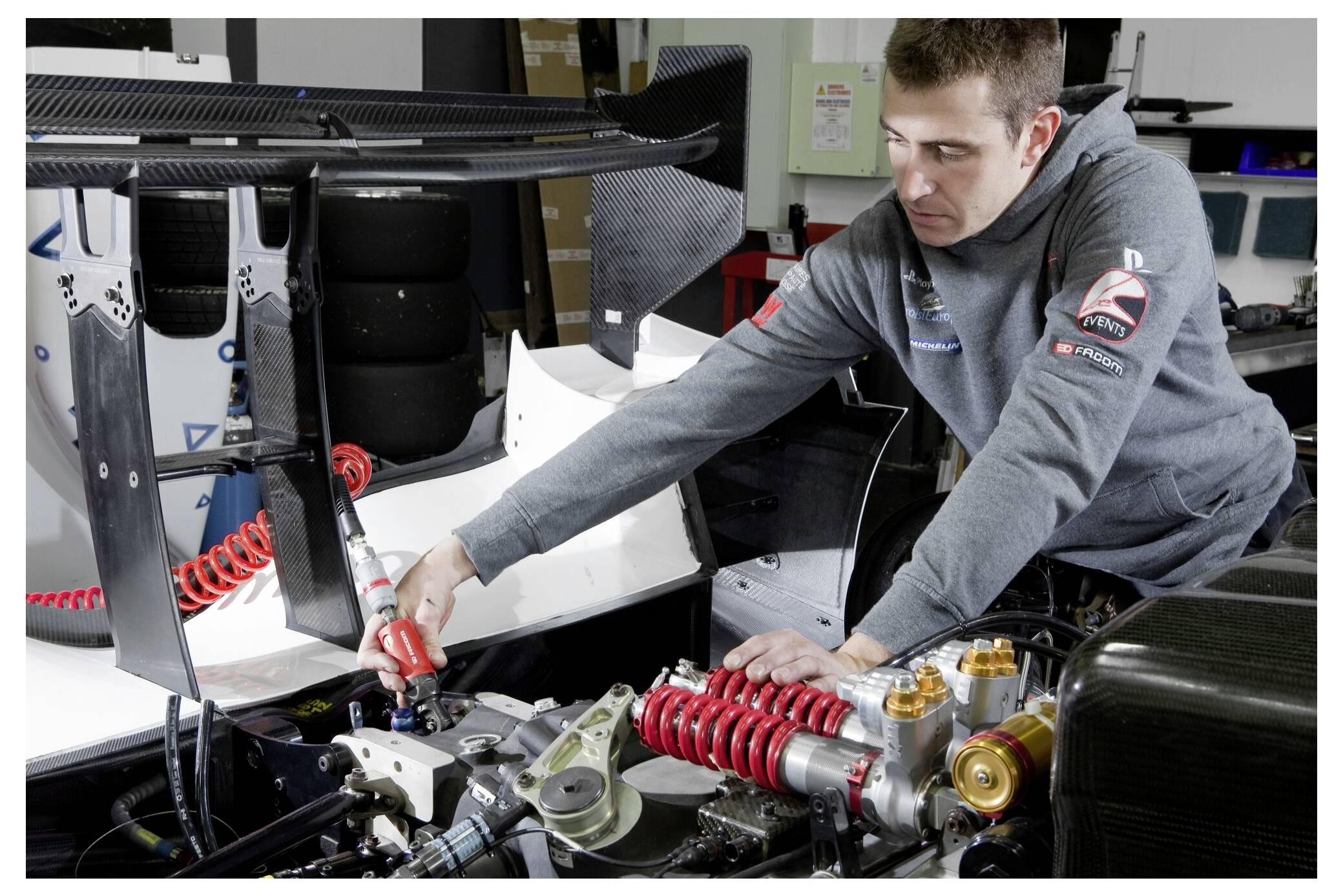 A mechanic works on a race car, adjusting components near the rear wing and suspension, surrounded by tools and equipment in a garage.