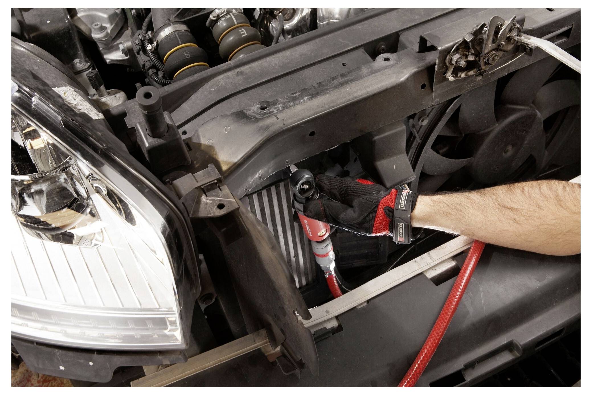 A person wearing a glove uses a tool to service an open car engine, highlighting a repair or maintenance activity.