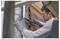 A person in protective gloves and glasses is installing insulation material on a building exterior, cutting it precisely around a window.