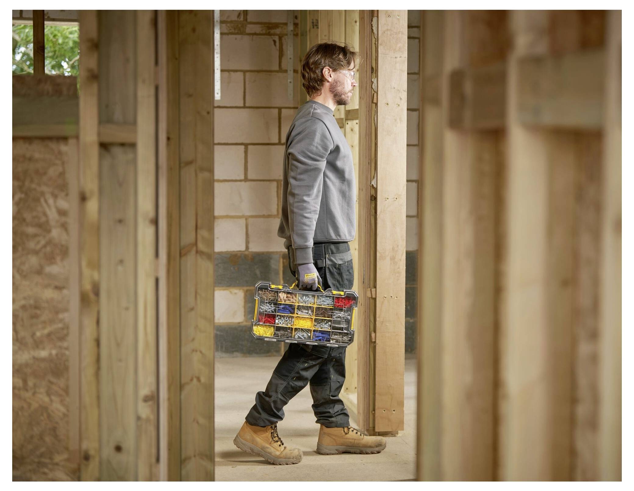 A person walks through a wooden-frame construction site holding a toolbox, wearing work gloves and boots.