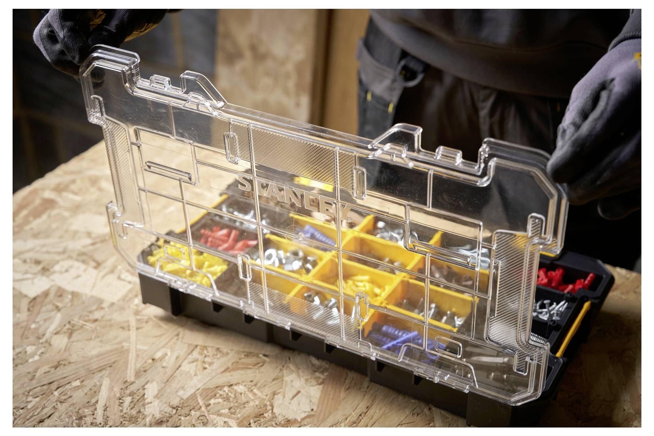 A person wearing gloves holds an open compartmentalized toolbox filled with various screws, bolts, and anchors, on a wooden surface.