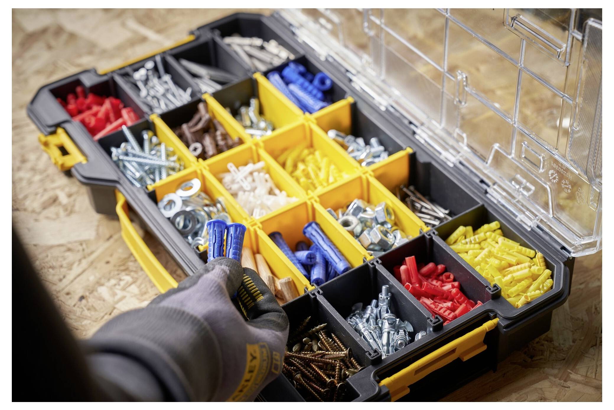 An open toolbox on a wooden surface filled with various screws, bolts, and fasteners in different compartments; a hand reaches for an item.