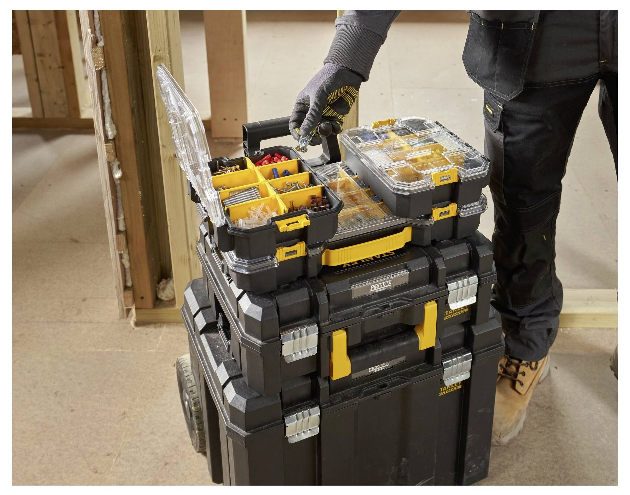 A person in workwear organizes tools in a large, stacked toolbox on wheels, inside a partially constructed wood-framed building.