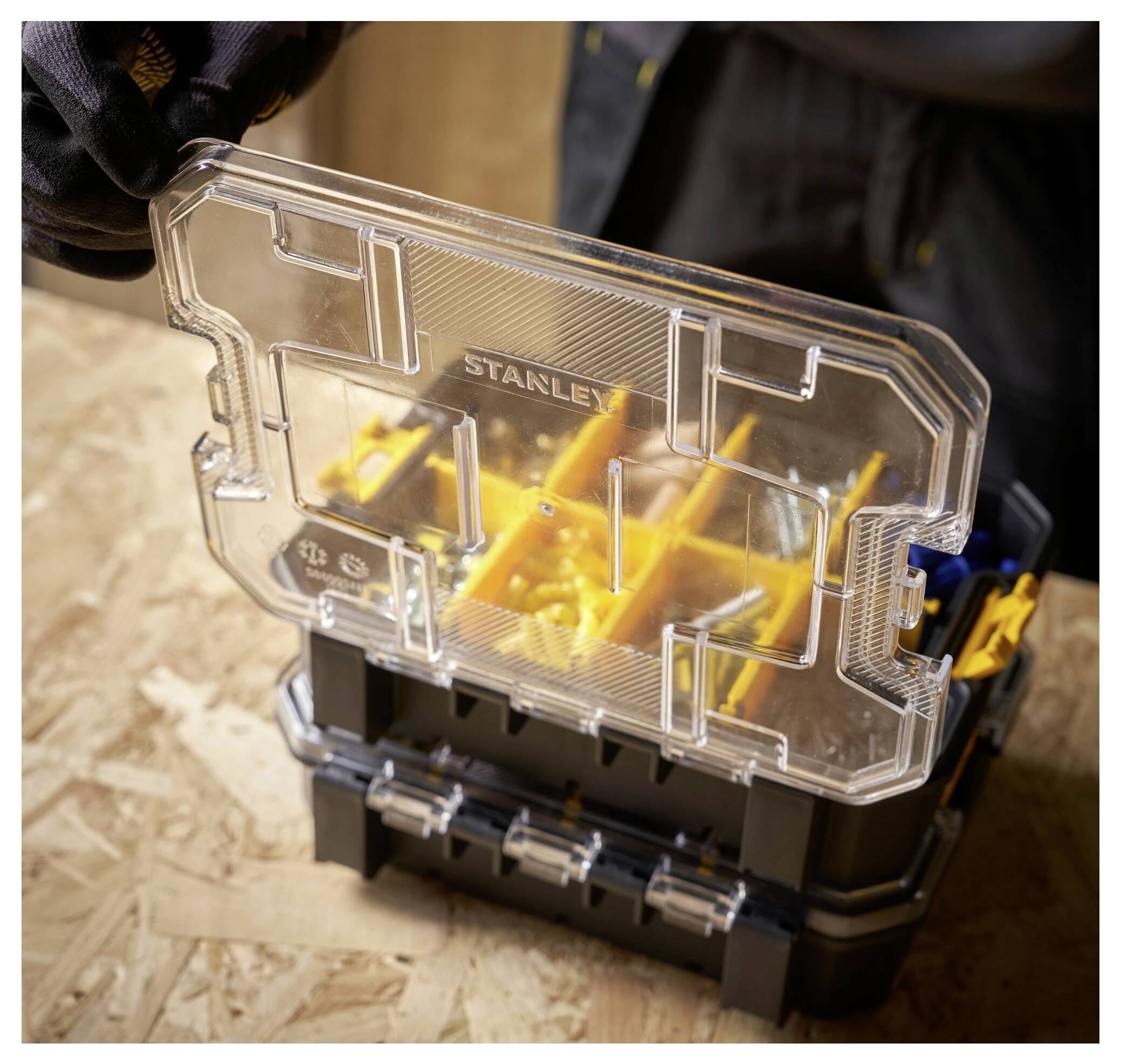 A person opens a black and yellow plastic toolbox with a transparent lid on a wooden surface.