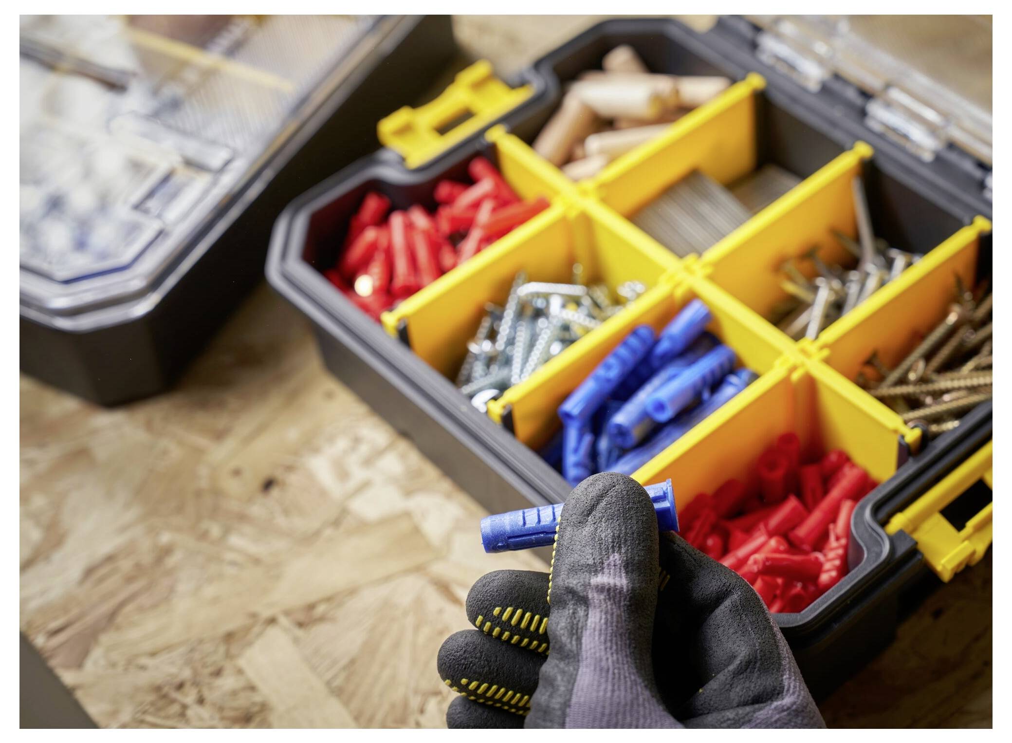A gloved hand holds a blue wall plug above an open toolbox organized with screws, plugs, and other small hardware on a wooden surface.
