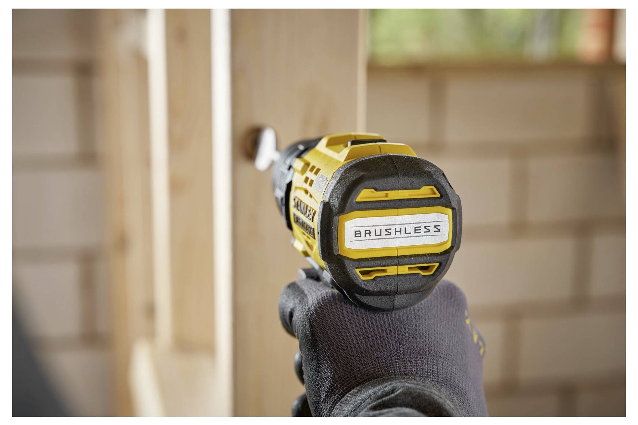 A gloved hand holds a yellow cordless brushless drill, driving a screw into a wooden beam in a construction environment.