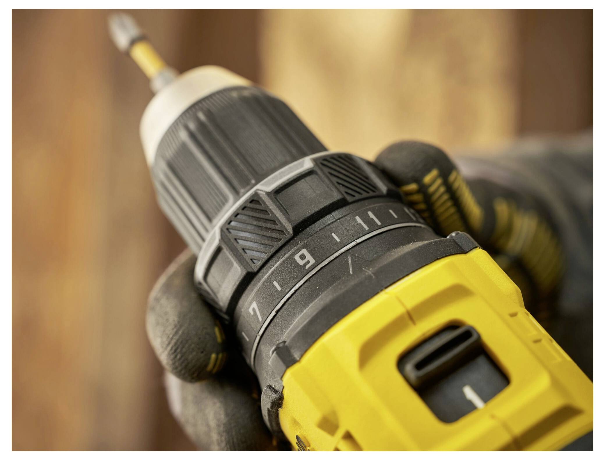 A person wearing gloves holds a yellow and black power drill, aimed forward, with a focus on the drill's adjustable settings.