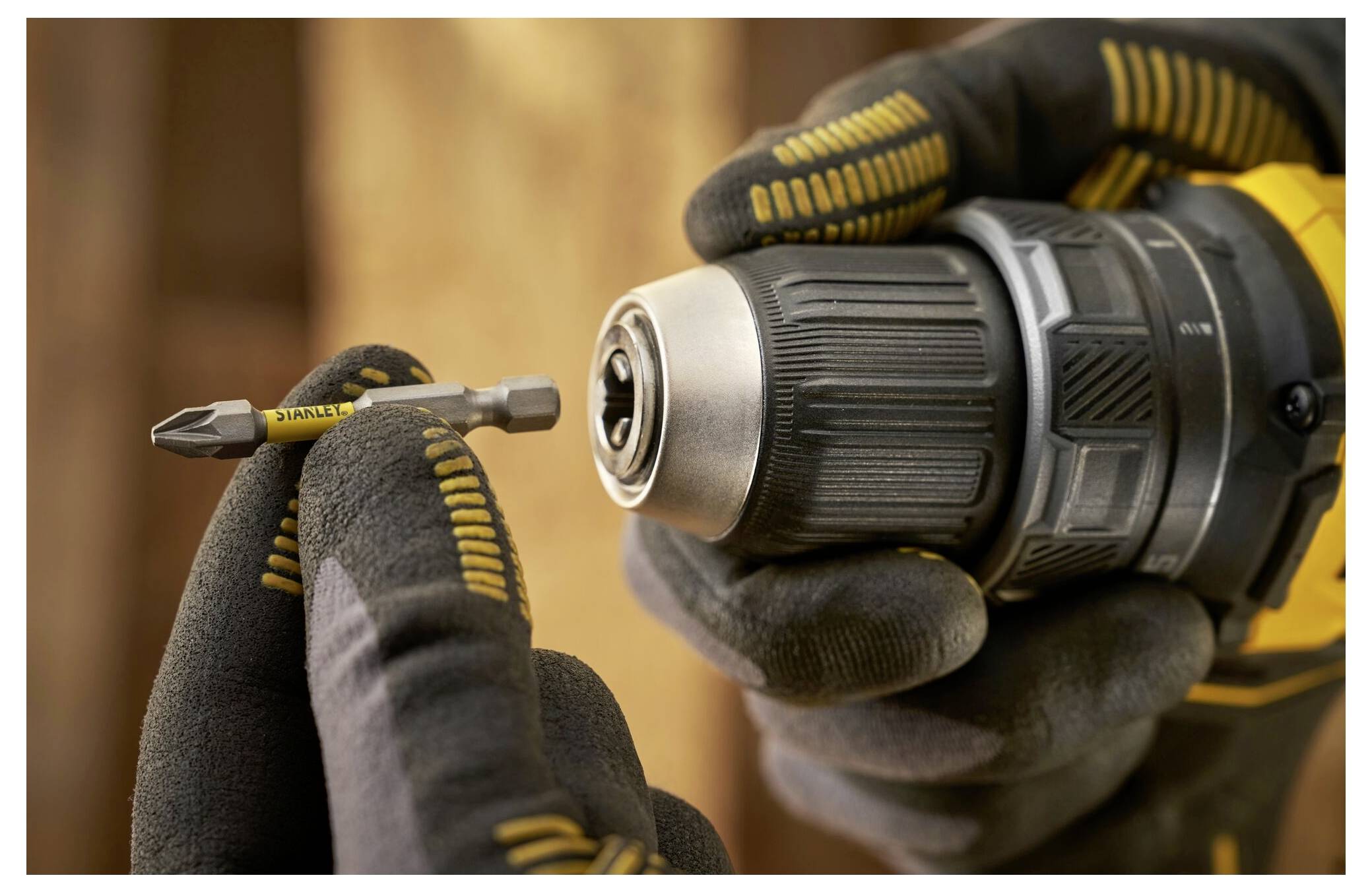 A person wearing protective gloves holds a drill in one hand and a drill bit in the other, about to insert the bit into the drill.