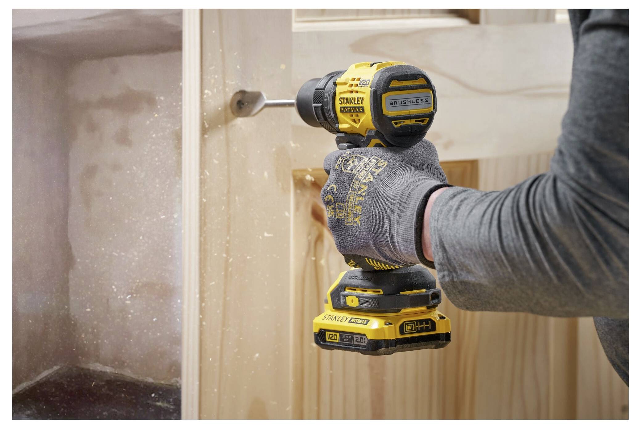 A person wearing gloves uses a yellow and black drill on a wooden surface indoors, highlighting woodworking or construction activity.