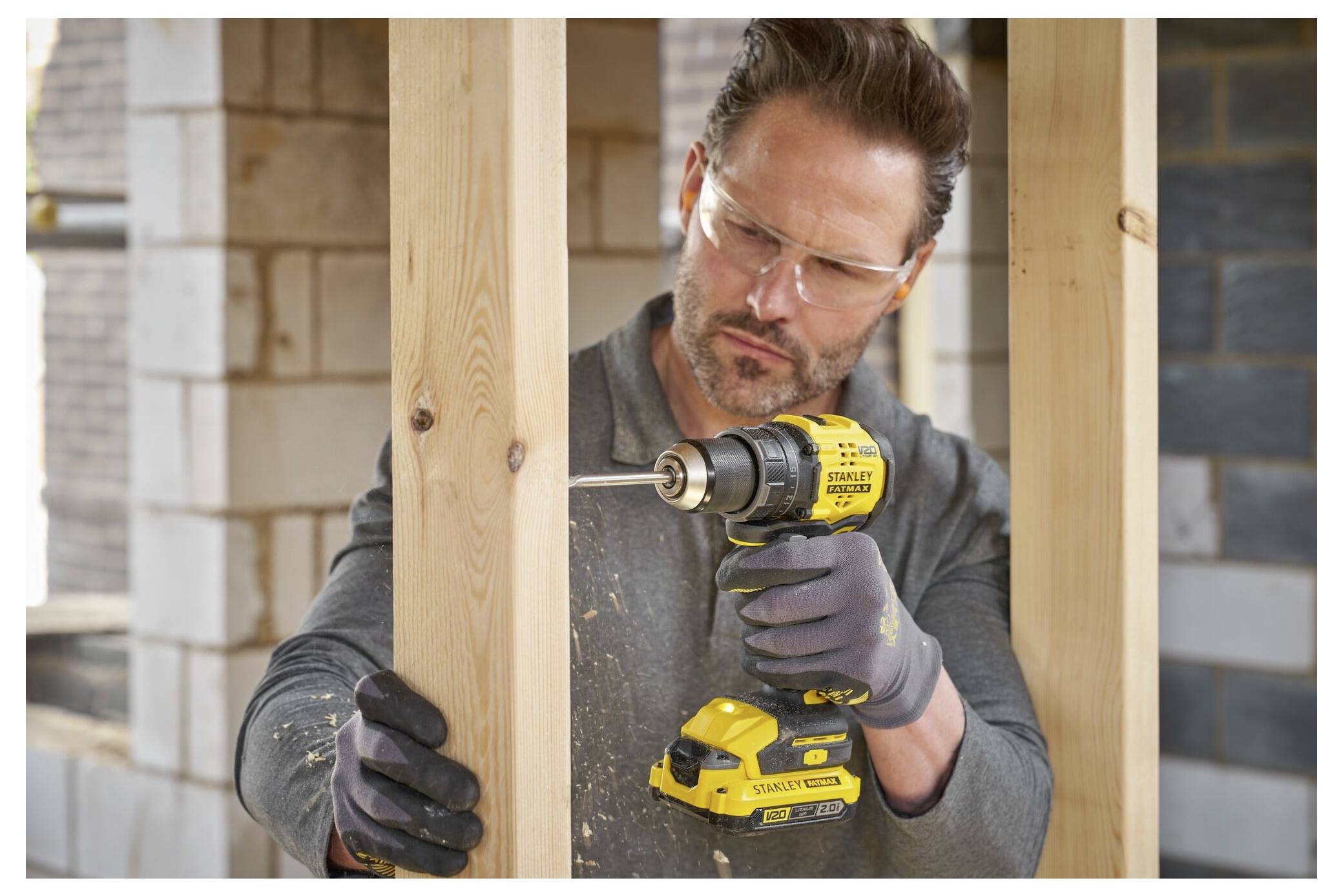 A person wearing safety goggles and gloves uses a yellow power drill to make a hole in a wooden beam on a construction site.