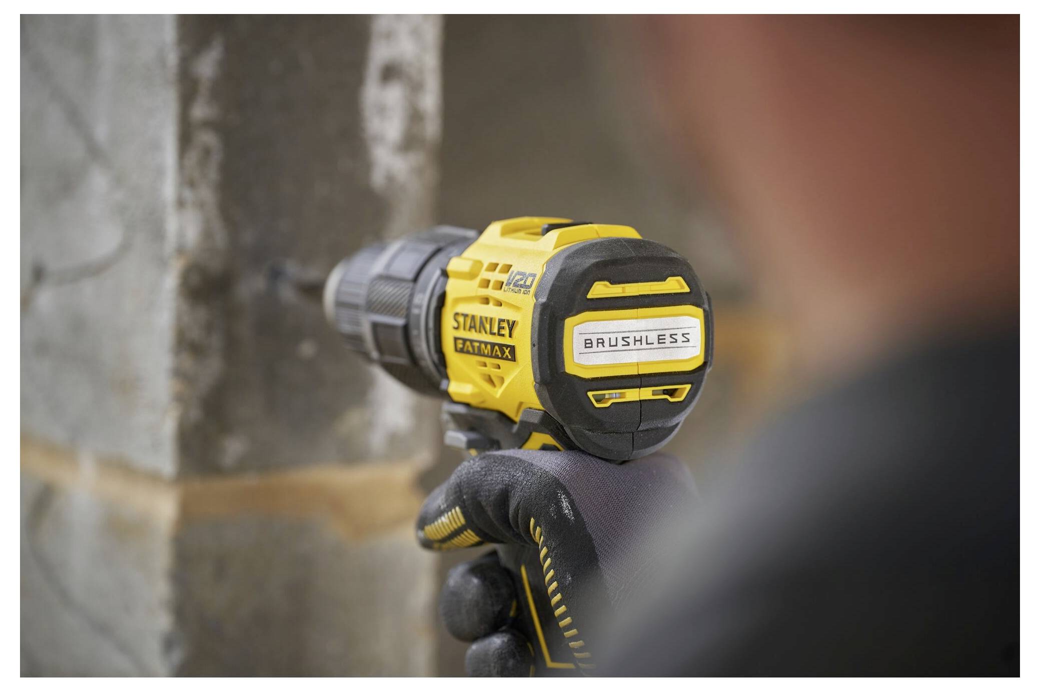 A person uses a yellow 'Stanley Fatmax Brushless' drill to make a hole in a concrete wall, focusing on the tool's rear label.