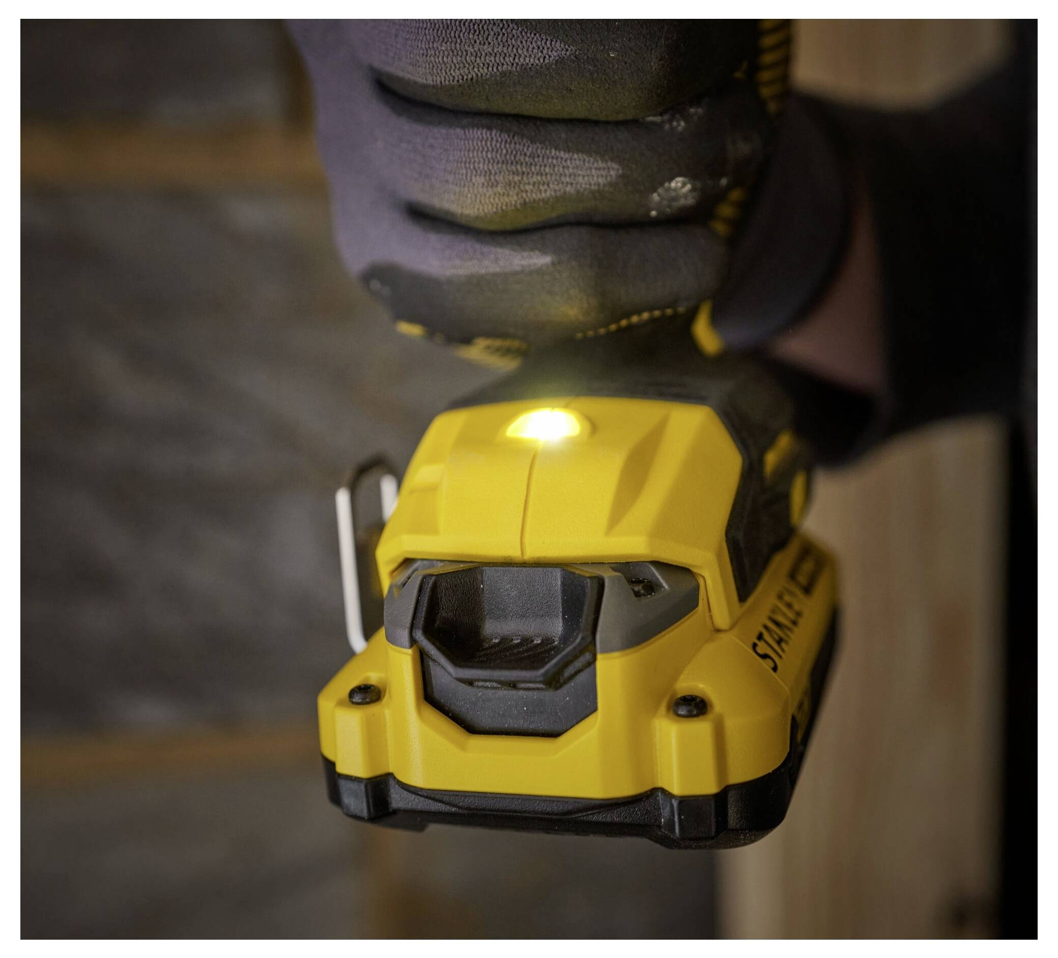 A person wearing gloves holds a yellow handheld power tool, likely a drill, with a bright LED light, against a wooden surface.