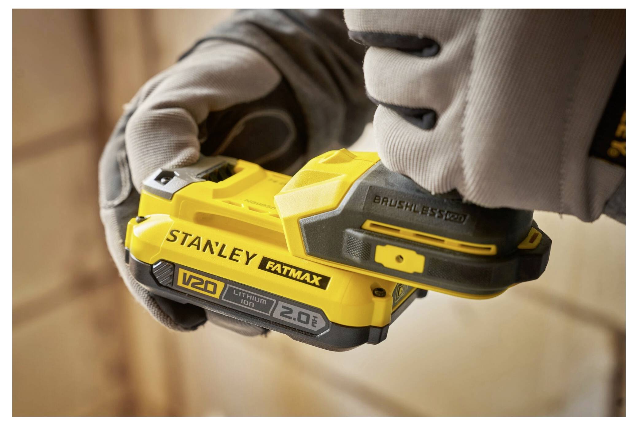 A person wearing gloves is holding a yellow Stanley Fatmax 20V drill, focusing on the battery pack labeled 'V20 Lithium Ion 2.0 Ah'.