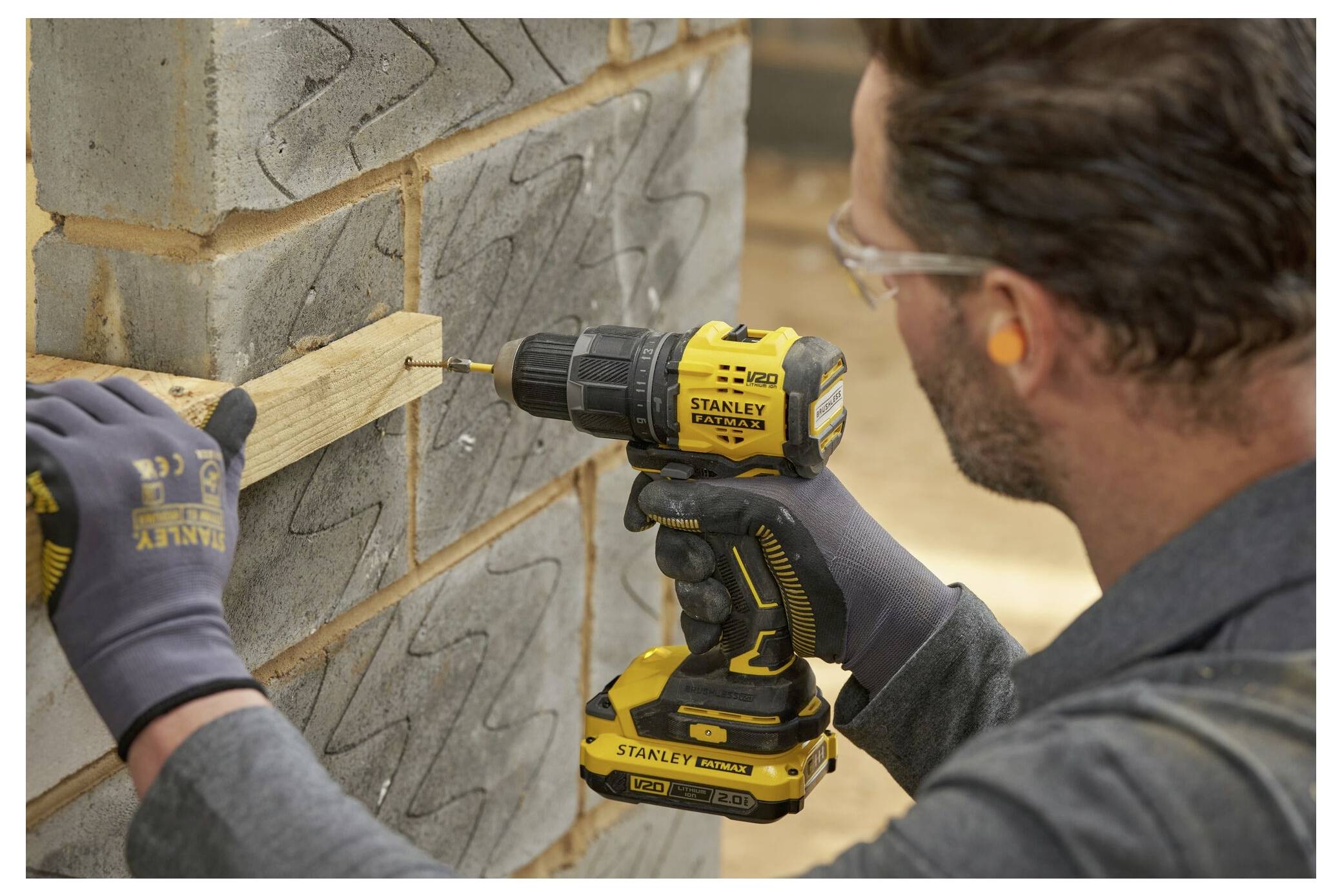 A person uses a yellow cordless drill to secure a wooden plank to a block wall. They wear gloves, safety glasses, and ear protection.