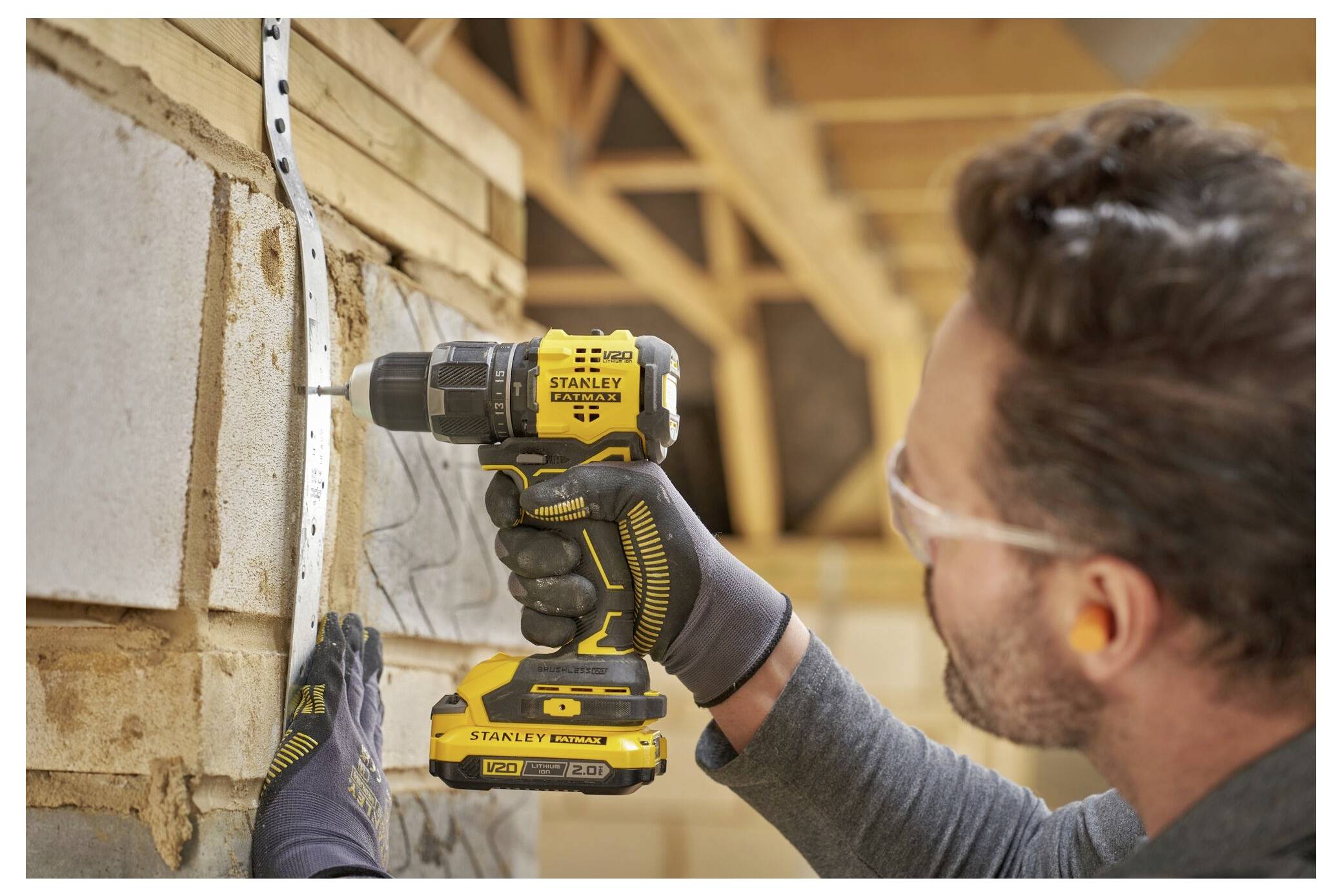 A person wearing safety goggles uses a yellow power drill to secure a metal bracket onto a wooden structure inside a building under construction.