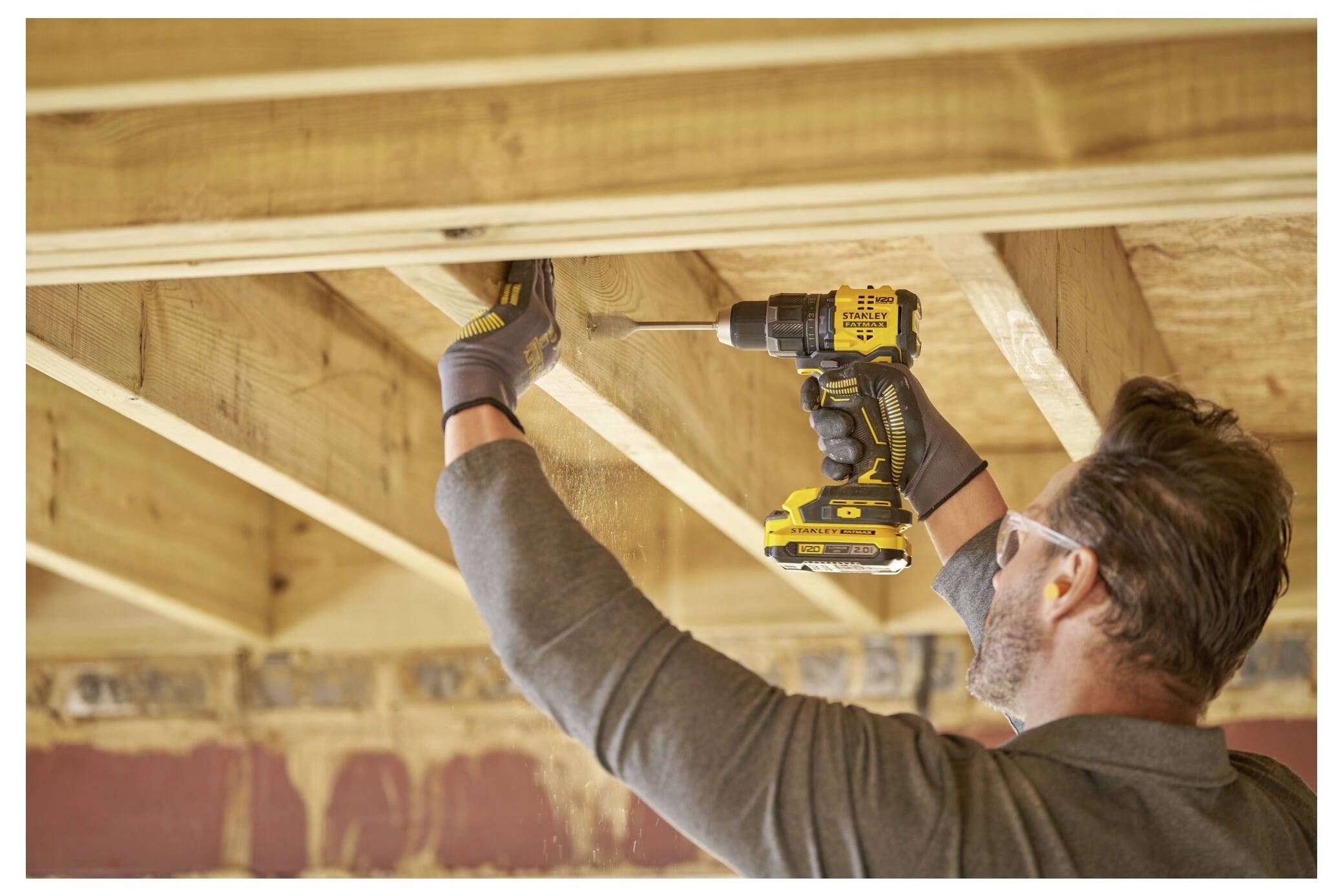 A person wearing gloves and goggles is using a yellow power drill to attach a wooden beam in a partially constructed ceiling.