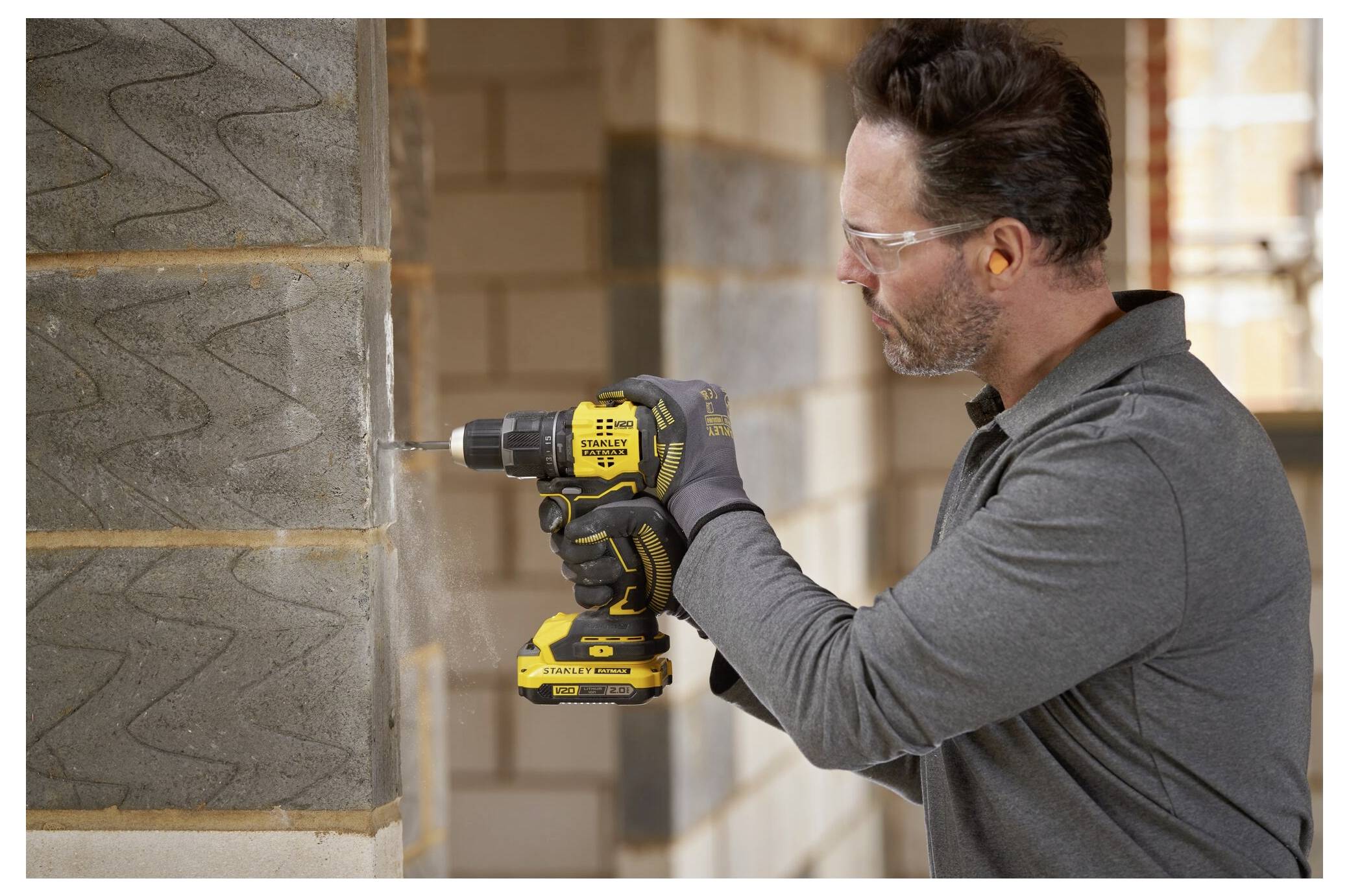 A person in protective gear uses a cordless drill to make a hole in a concrete wall inside a partially constructed brick building.