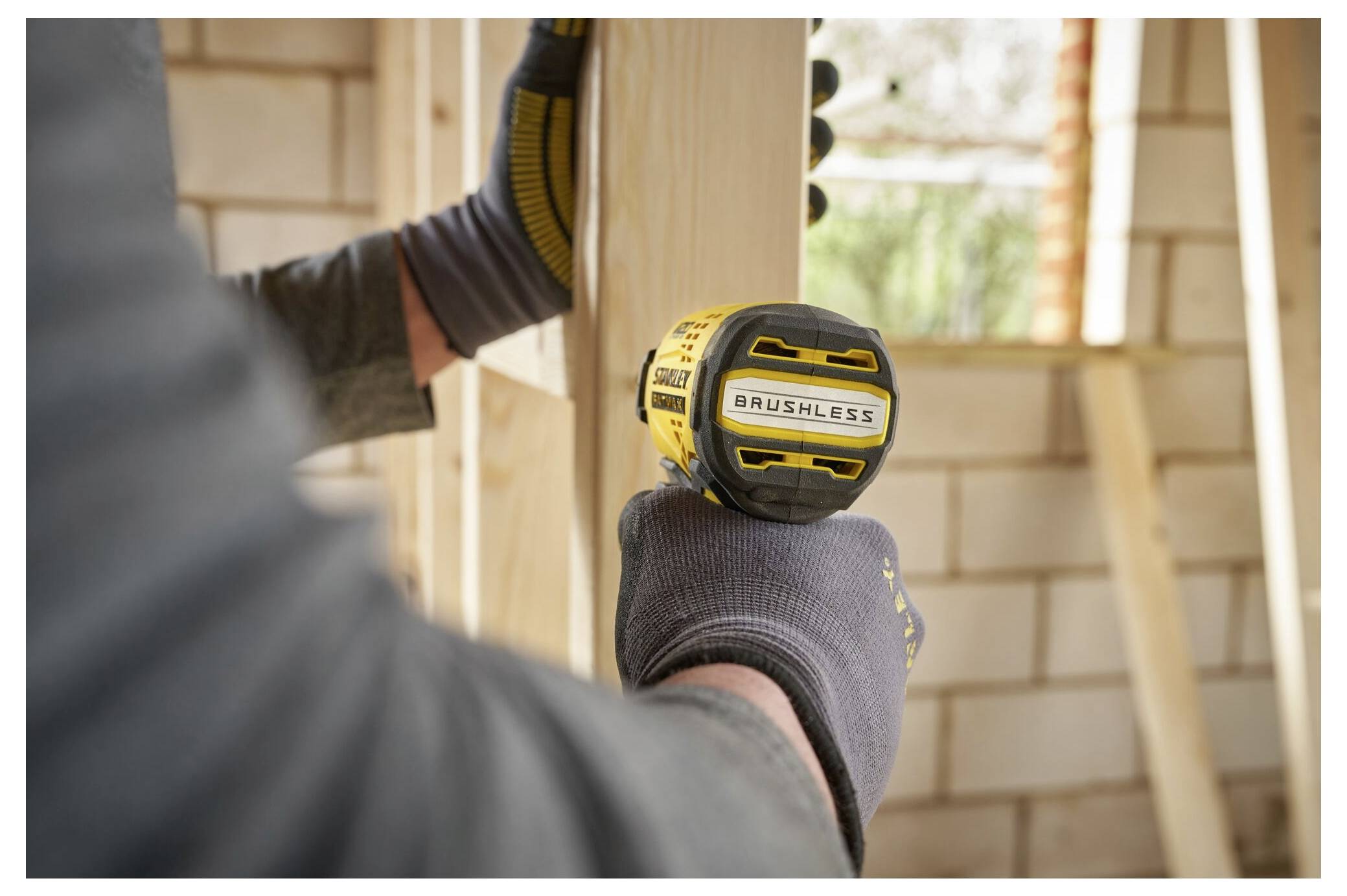 A person wearing gloves uses a yellow and black brushless drill on a wooden frame, with a partially completed brick wall in the background.