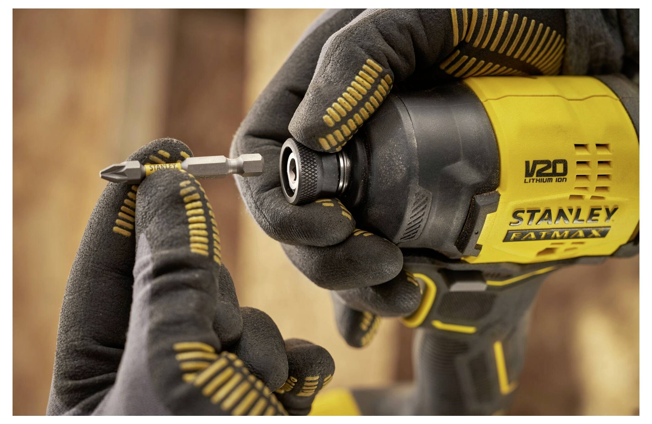 'Close-up of hands wearing gloves holding a drill, inserting a drill bit. The drill is a yellow V20 Stanley Fatmax Lithium Ion.'