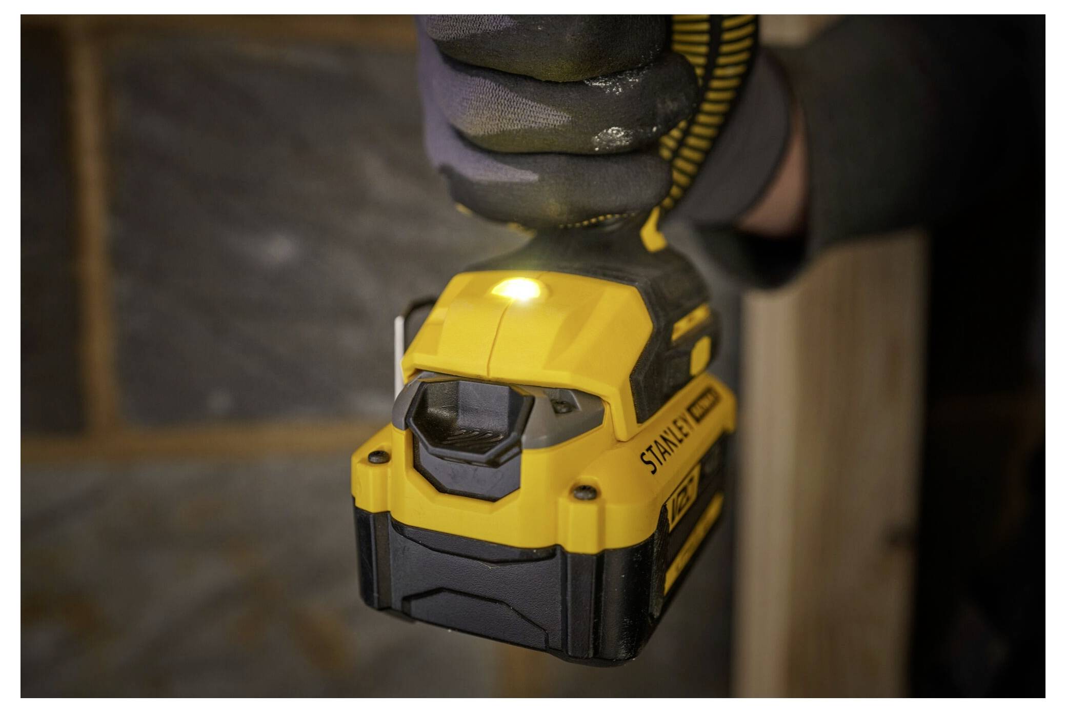 A person holds a yellow cordless power tool labeled 'Stanley'. The tool is used in a construction setting, with gray stone walls visible.