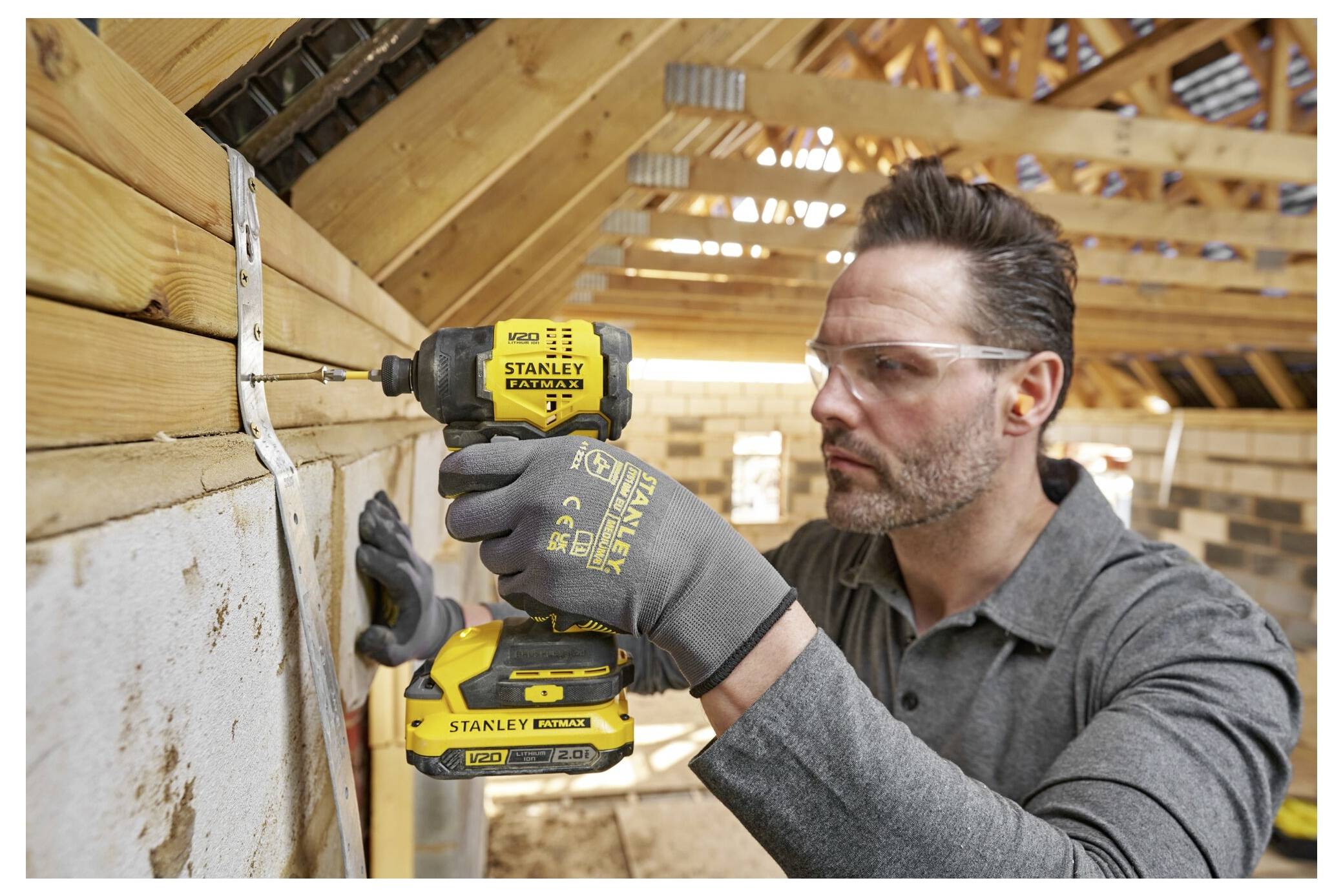 A man wearing safety goggles and gloves uses a power drill to install wooden beams in a house under construction.