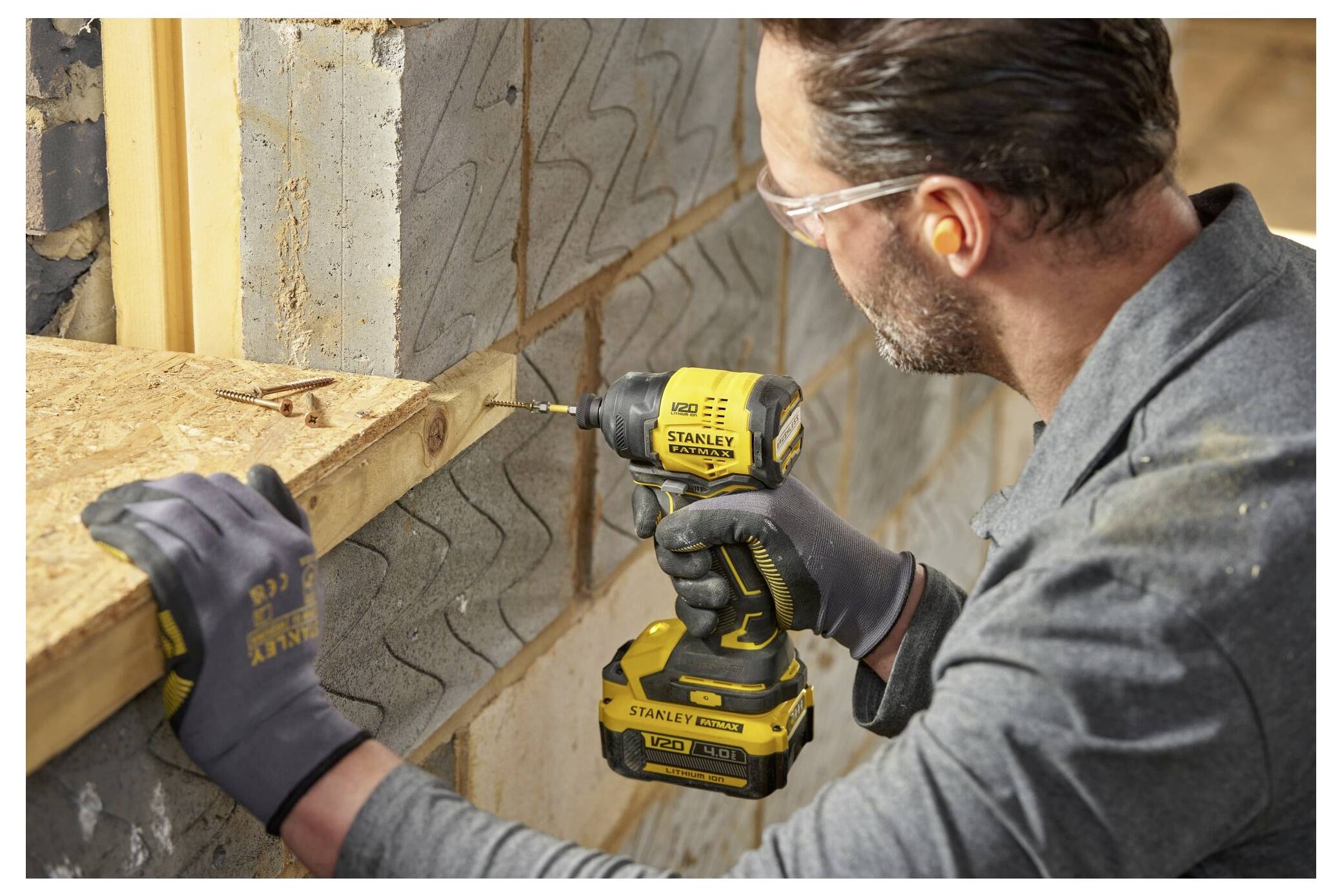 A person wearing safety gear uses a yellow power drill to screw wood planks together in a construction setting.