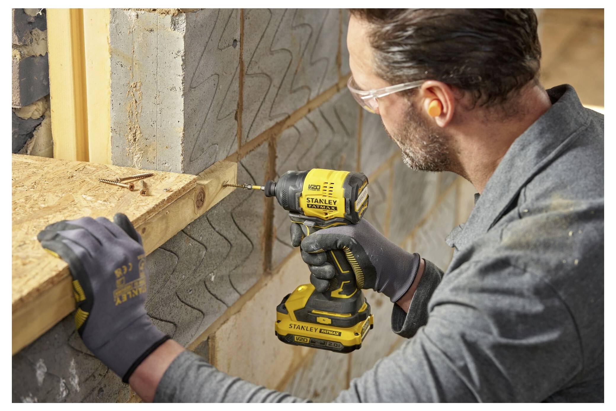 A person using a yellow and black power drill to secure wood against a block wall in a construction setting, wearing protective gear.
