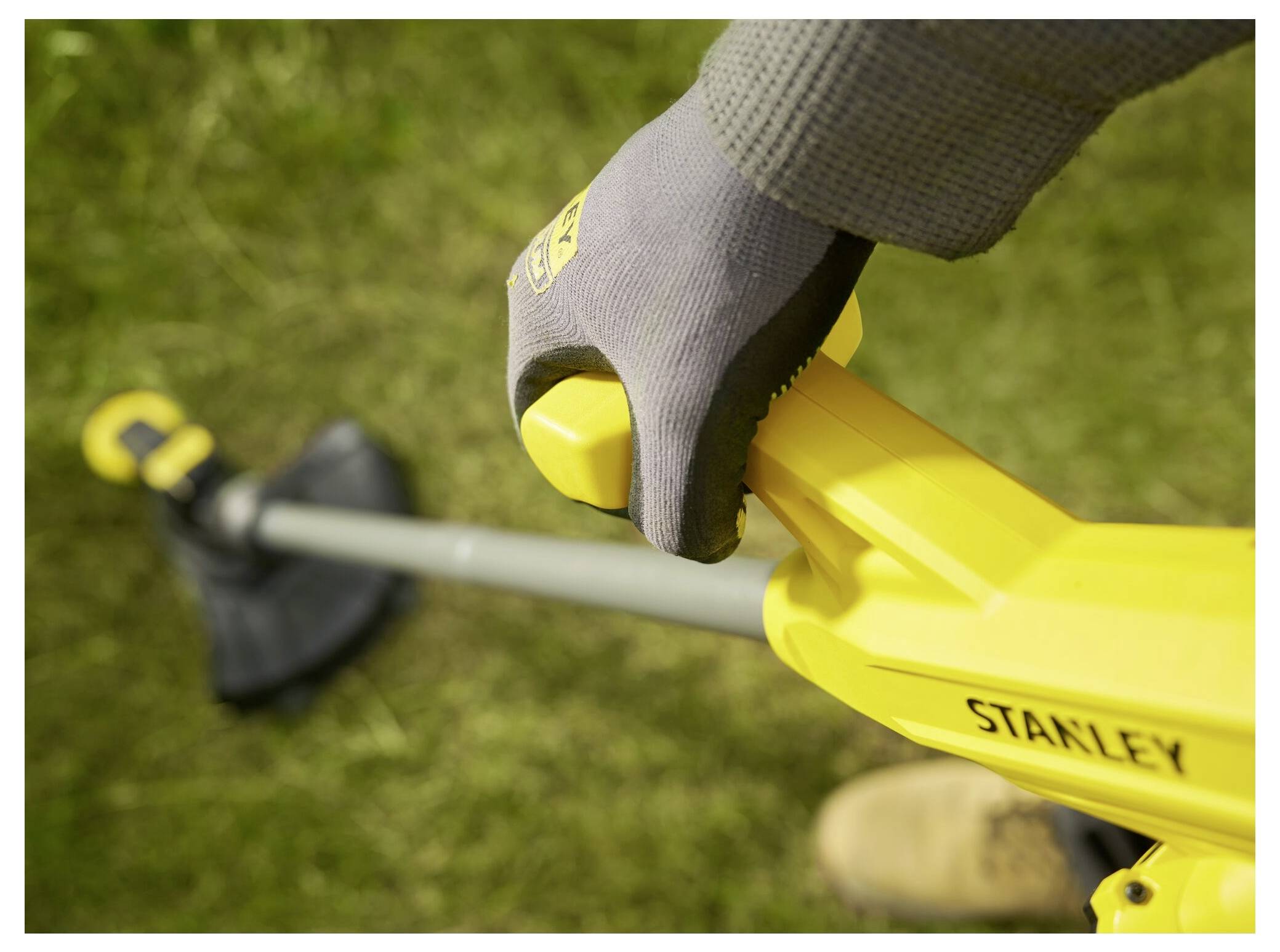 A person wearing gloves holds the handle of a yellow grass trimmer on a grassy area.