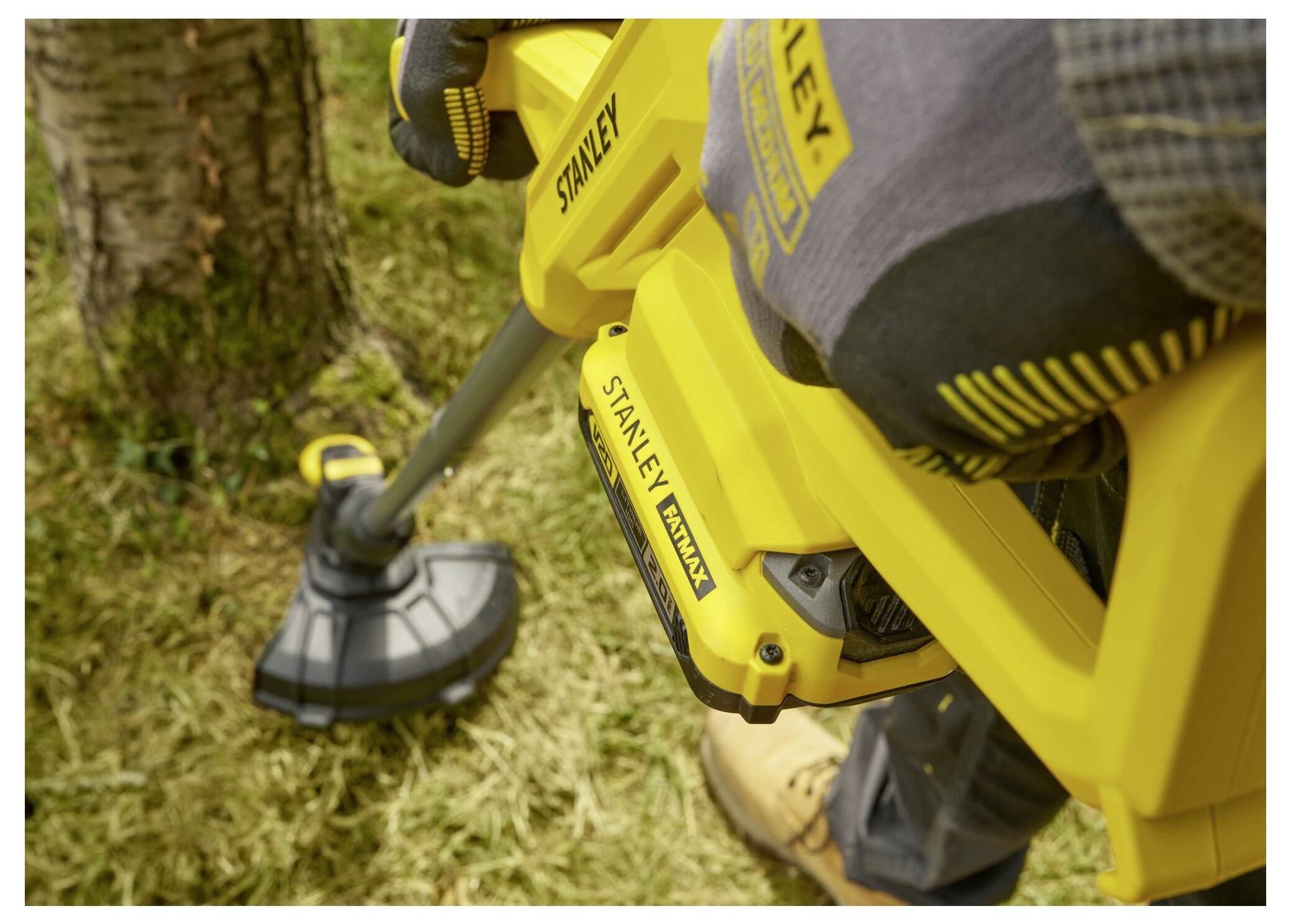 'A person uses a yellow Stanley grass trimmer to cut grass around a tree, wearing gloves and boots for protection.'