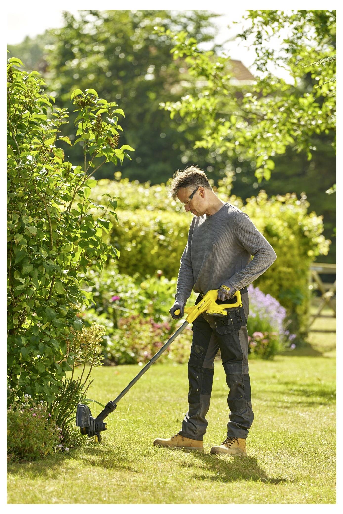 A person using a yellow cordless trimmer to cut grass near a bush in a sunny garden. The background features lush greenery.