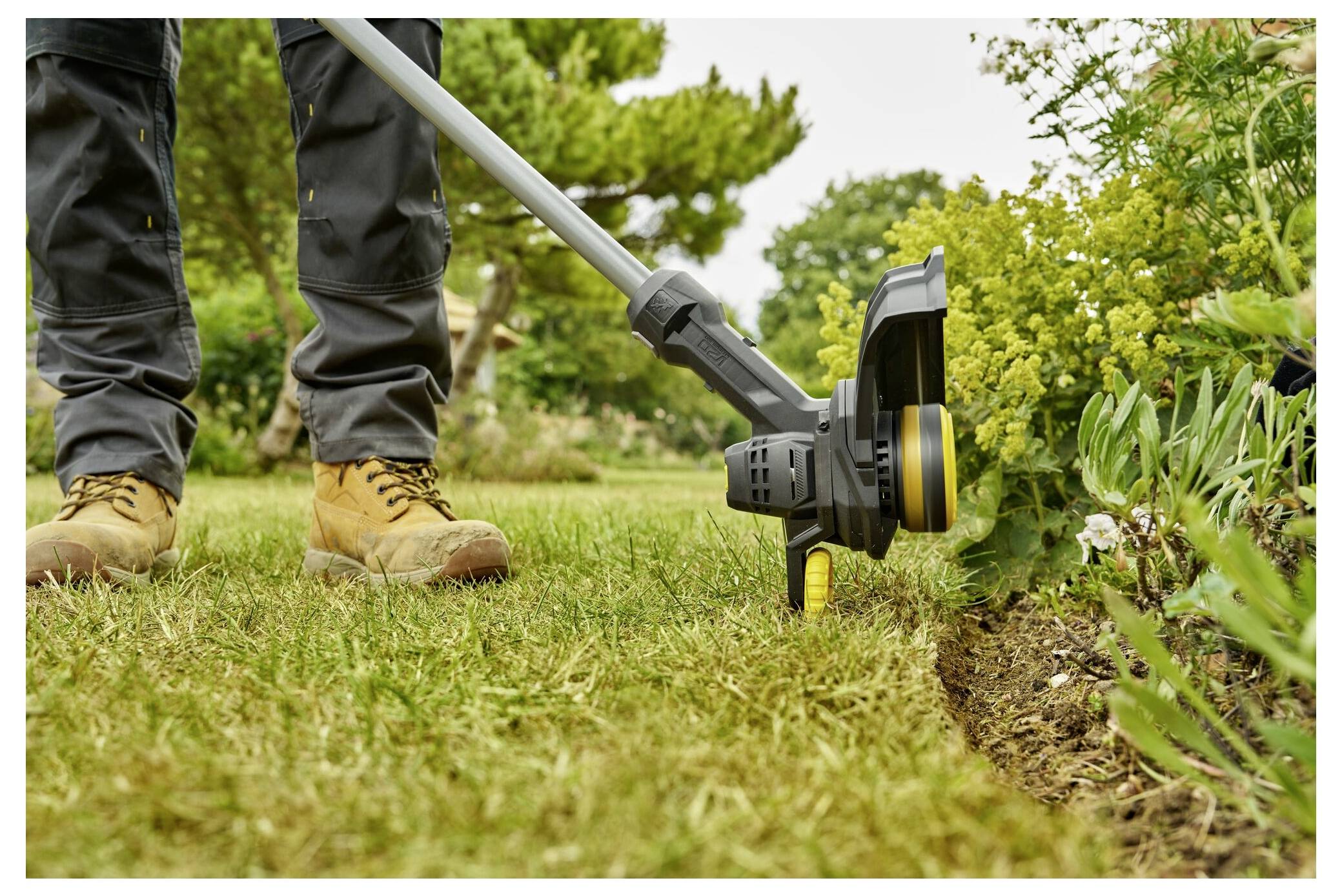 A person is using a string trimmer to edge a garden. They wear protective boots and pants. Lush green plants and grass surround the area.
