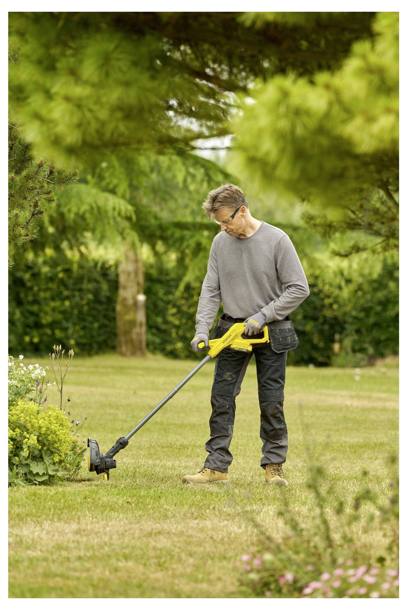 A person trims the grass using a yellow string trimmer in a garden surrounded by trees and shrubs.