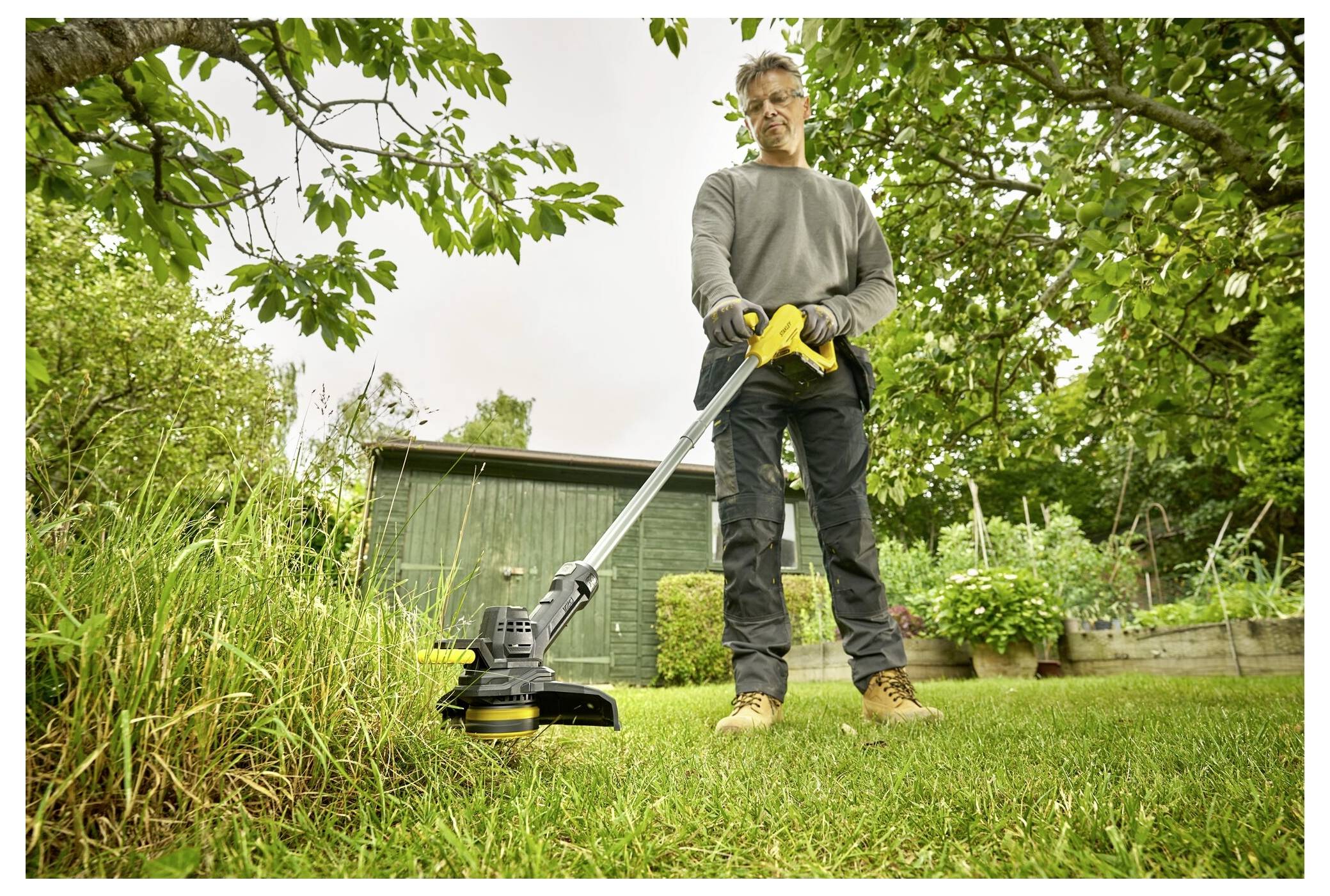 A person uses a grass trimmer to cut the lawn in a garden with trees and a small shed in the background, wearing protective gloves.